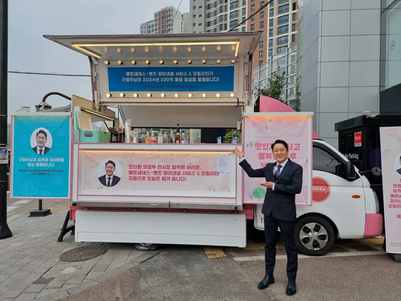 구명우 딜러의 얼굴이 실린 배너가 걸린 커피차와 정장 차림의 남성이 있는 행사 현장